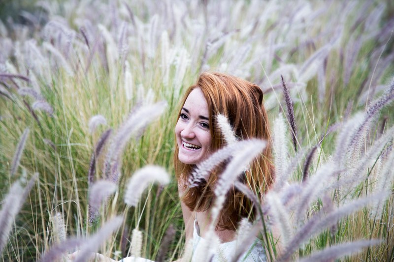 a woman sits laughing in a field of long purple tipped grasses, woman with purple grasses 4k