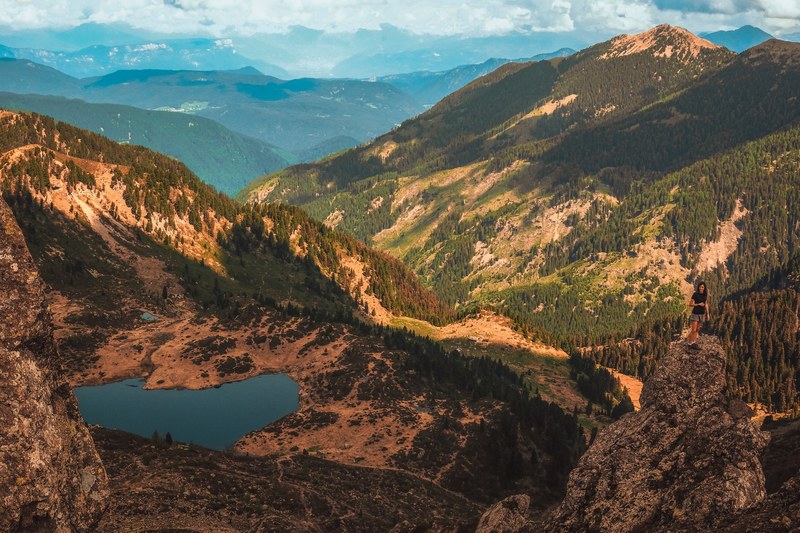 a woman standing on a ledge over a mountain green valley with a small lake, ledge over mountain valley 4k