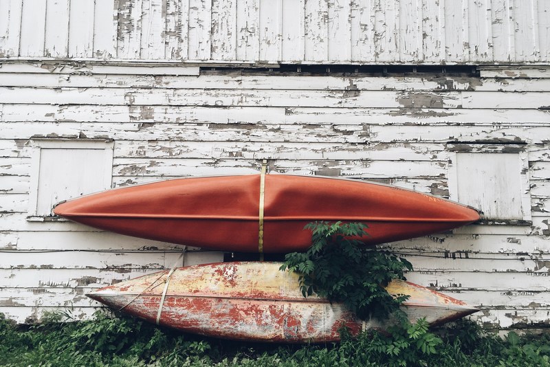 two used kayaks leaning on an old barn wall with peeling white paint and unkept grass, _two kayaks leaning on a wall 4k