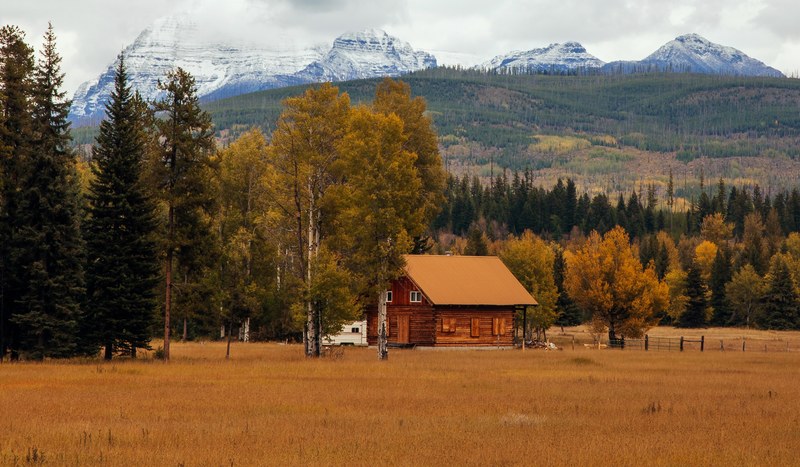 small log cabin house in glacier national park at the edge of a forest and field with snow capped mountains in the background, quiet cabin in the mountains 4k