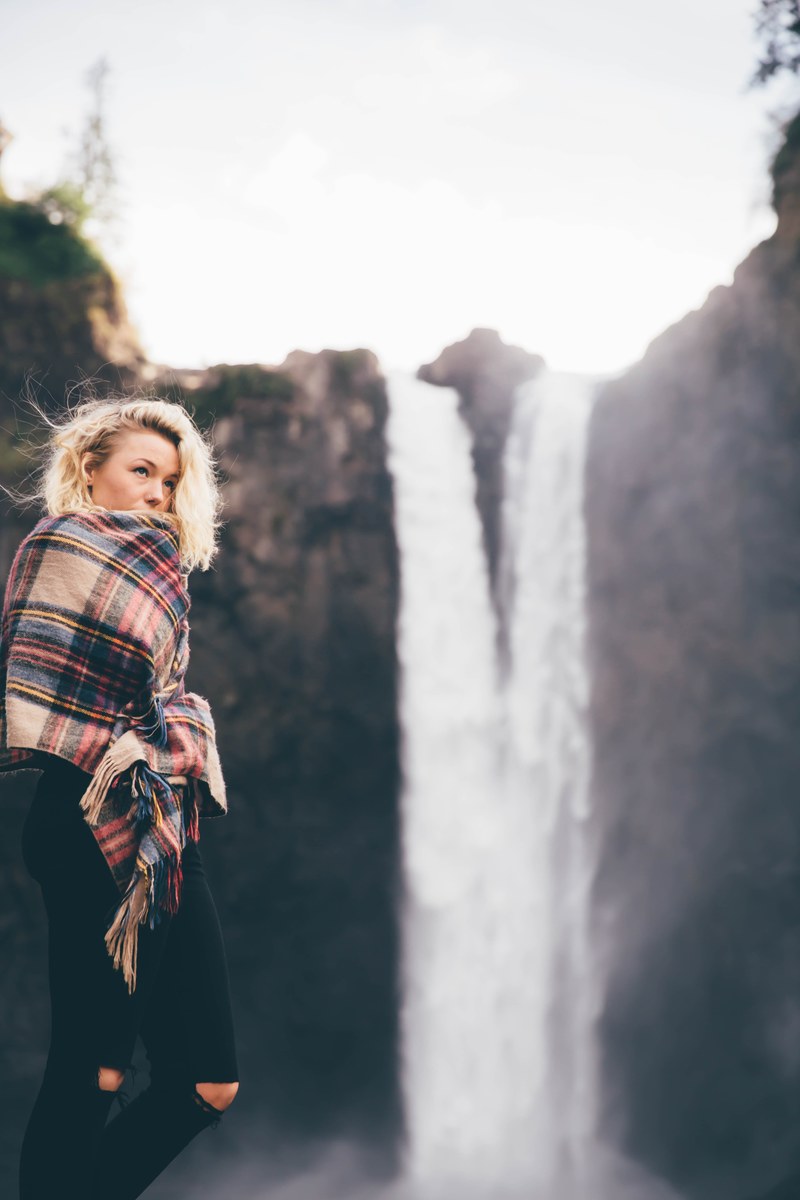 a woman wrapped in a plaid scarf or blanket standing on the edge of snoqualmie falls, woman by a waterfall 4k