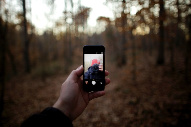 a young man holding out an iphone for a selfie alongside his camera in the woods, young man holding iphone and doing a selfie 4k
