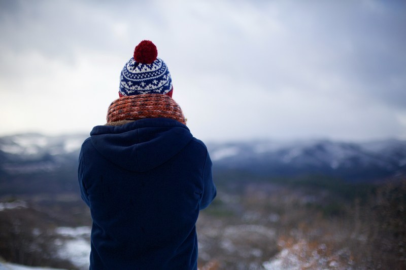 the back of a person with crossed arms wearing a knit cap scarf and blue coat is visible as he or she stares out into snowy wilderness, person stares into wilderness 4k