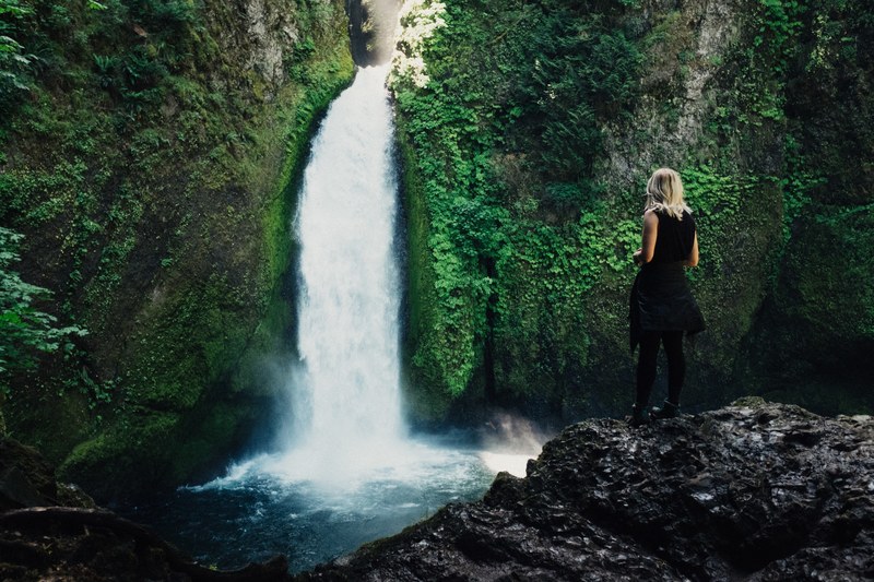 a blonde woman standing on a rock overlooking a pond with a waterfall tumbling into it, on a rock near a waterfall 4k