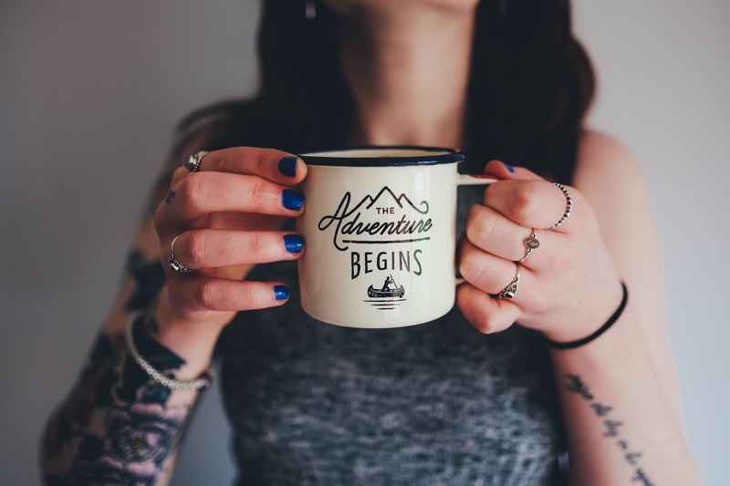 a woman with tattooed arms holding an enamel cup with the adventure begins written on it, the adventure begins cup 4k