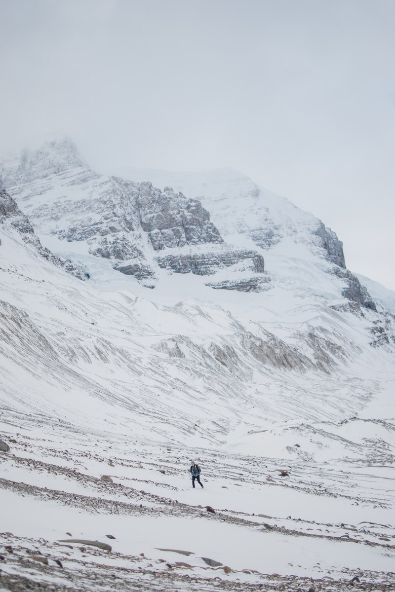 hiker in dark colors walks at the base of the snowy icy athabasca glacier, within titans 4k