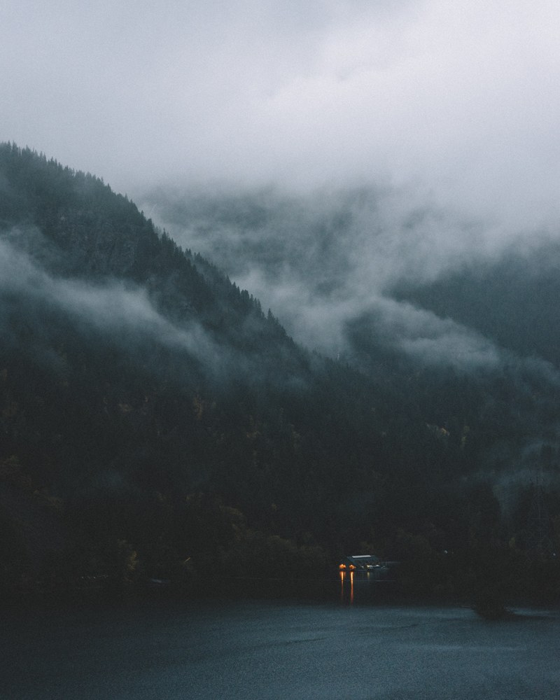 a dim shot of a cottage on the shore of a mountain lake with a thick fog on the slopes, quiet before the storm 4k