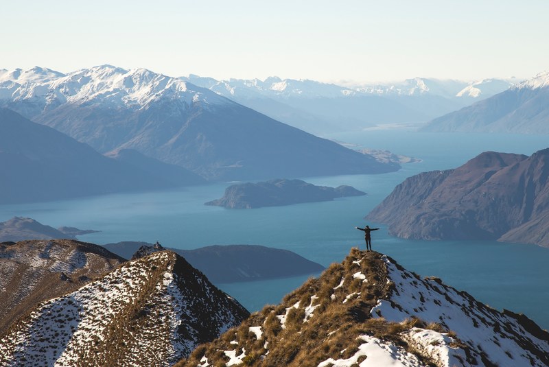 person stands on grassy snow covered summit overlooking water with arms outstretched, person on summit peak 4k