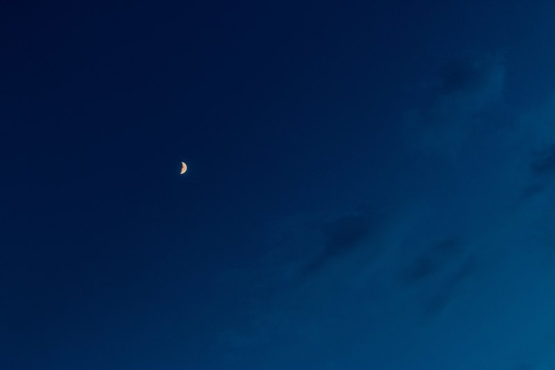 a crescent moon seen near fluffy clouds on an inky blue sky, crescent moon on a blue sky 4k