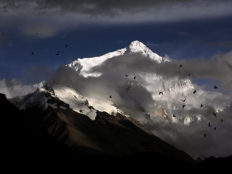 a tall snow capped mountain enveloped in gray clouds with birds flocking nearby, qomolangma 4k