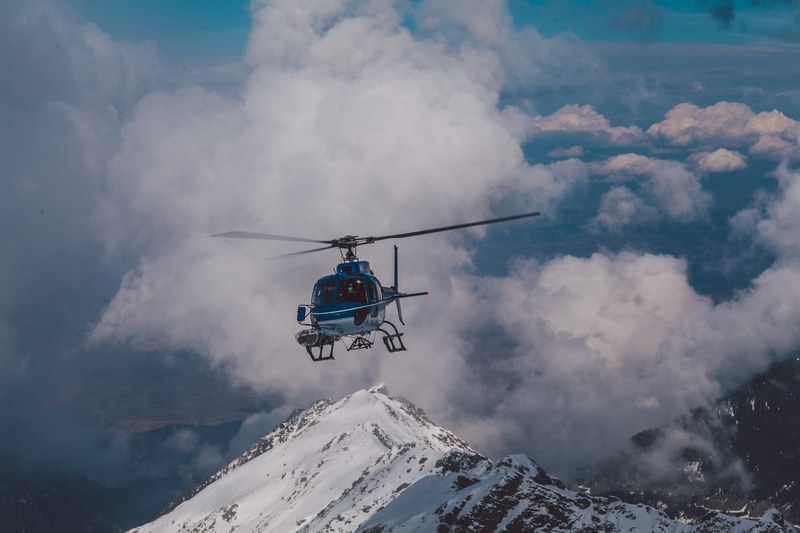 a blue helicopter is flying away from clouds and a snowcapped mountain, helicopter over mountain range 4k