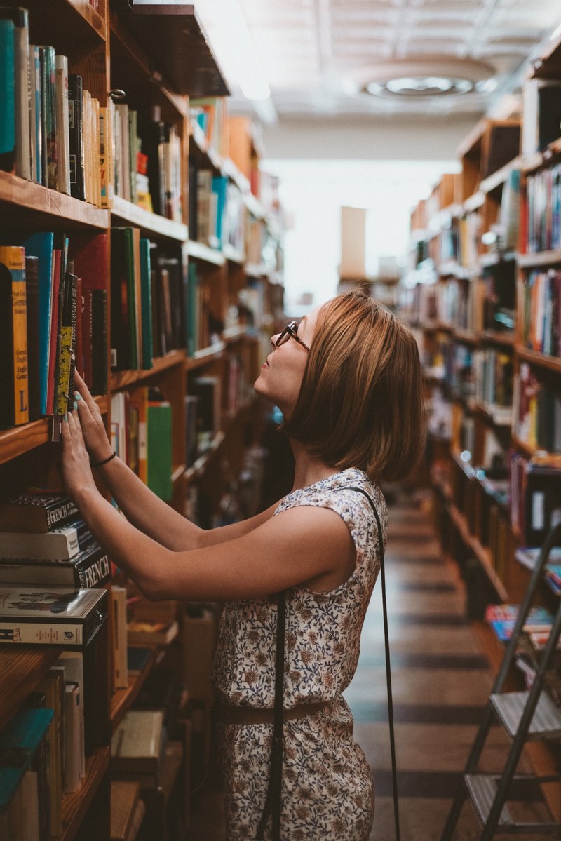 woman with short hair and glasses looking for a book in the shelves of a library in charlotte, lose yourself 4k
