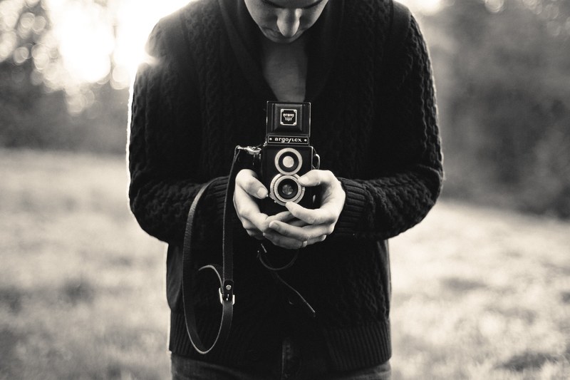 black and white shot of man in black cardigan holding vintage camera with grass in background, man holds vintage camera 4k