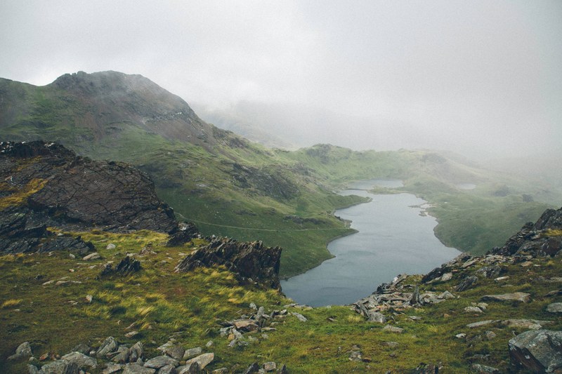 rocks line a grassy hillside near a lake of a foggy landscape, hillside vistas 4k