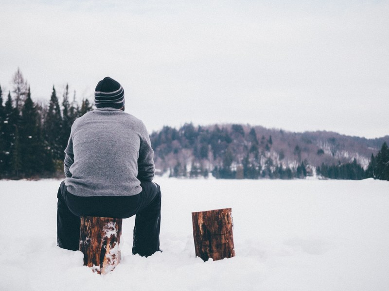 a man wearing a knit cap and gray sweater looks out into a snowy forest while sitting on a log, man sits on log in snow 4k