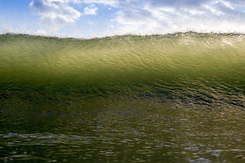 a perspective shot of a large wave about to roll over the sea beneath a blue cloudy sky, green wave under a blue sky 4k