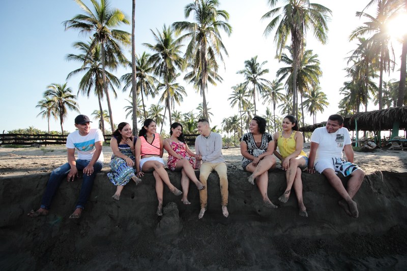 group of smiling friends sits on a stone wall at the sunny beach near palm trees, friends on wall at the beach 4k