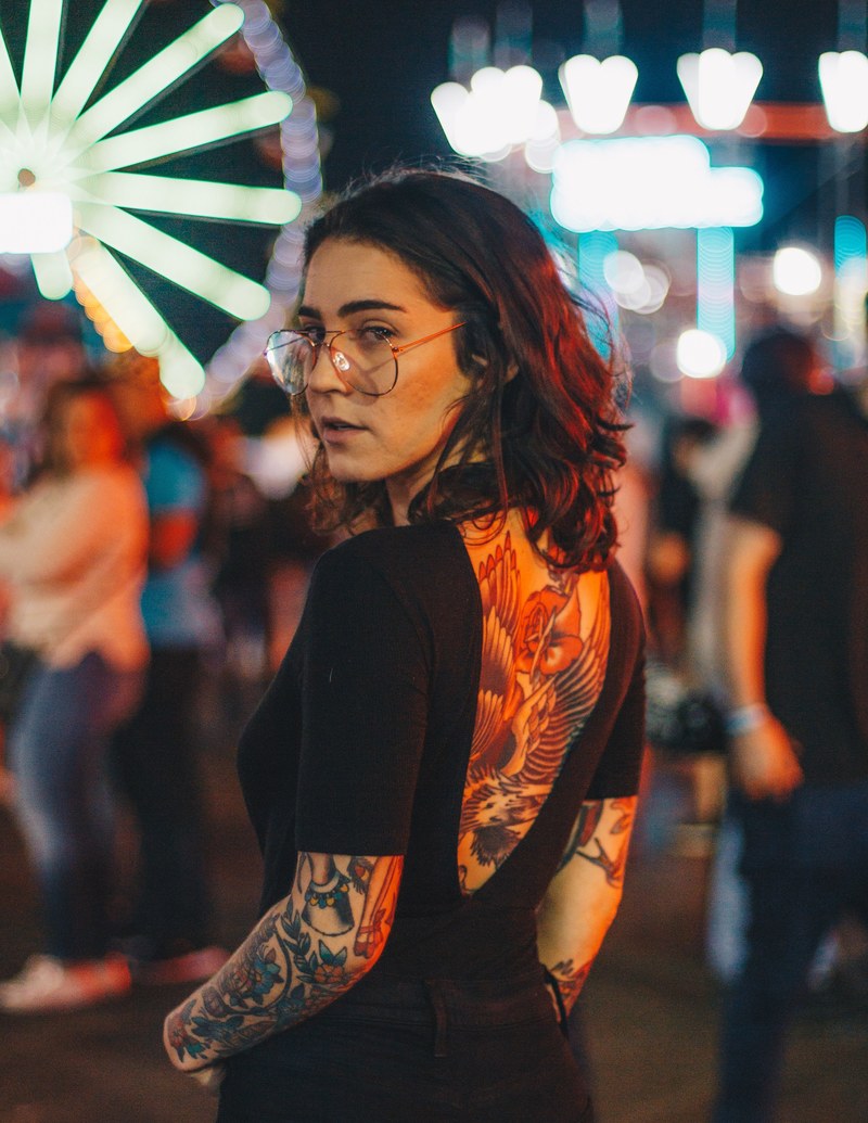 woman with the back and arm tattooed and glasses posing near amusement park at palm beach, fair 4k