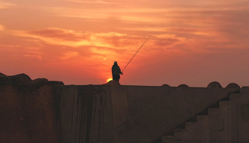 a silhouette of a fisherman standing atop a wall in asilah morocco during sunset, fishing in asilah at sunset 4k