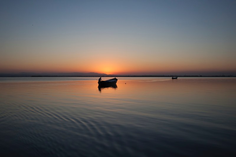 rowing boats sit on a tranquil lake in silhouette against a low sunset on the horizon, rowing boats at sunset_ 4k