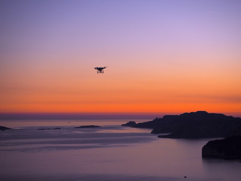 a drone hovering over the water and rocks of la ciotat with an orange and purple sunset in the background, drone over the water and rocks 4k