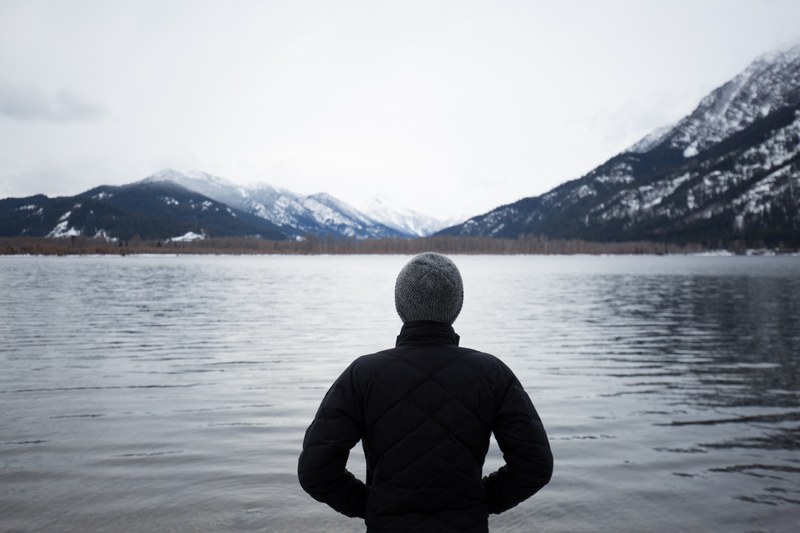 a person wearing a knit hat and puffy jacket looking out at lake wenatchee, man looking at water 4k