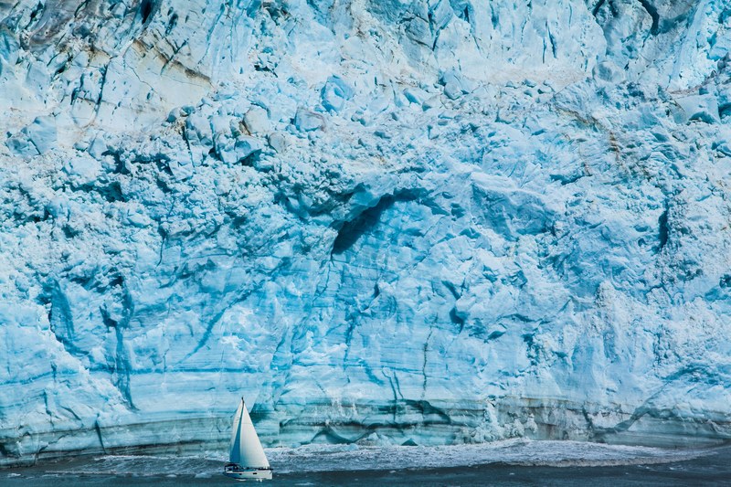 a steep light blue iceberg with a sailboat on the water below, high ice blue iceberg 4k
