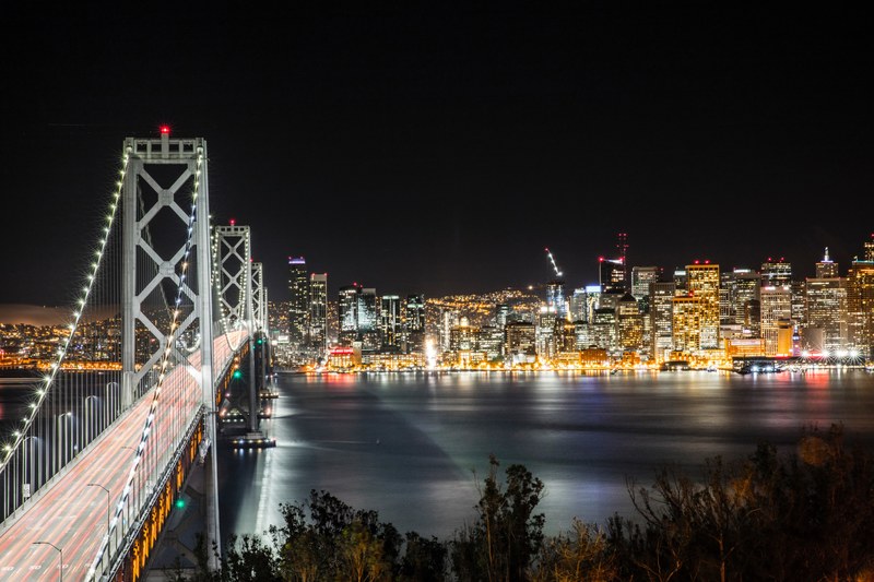 a long exposure shot of a bridge with red light trails on it and san francisco skyline on the other side, light rails on a san francisco bridge 4k