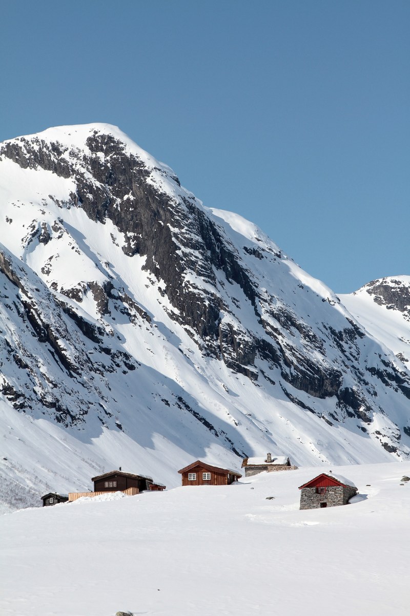 houses at the top of a hill with snow capped mountains in the background and blue skies, strynefjellets snowy cabins 4k