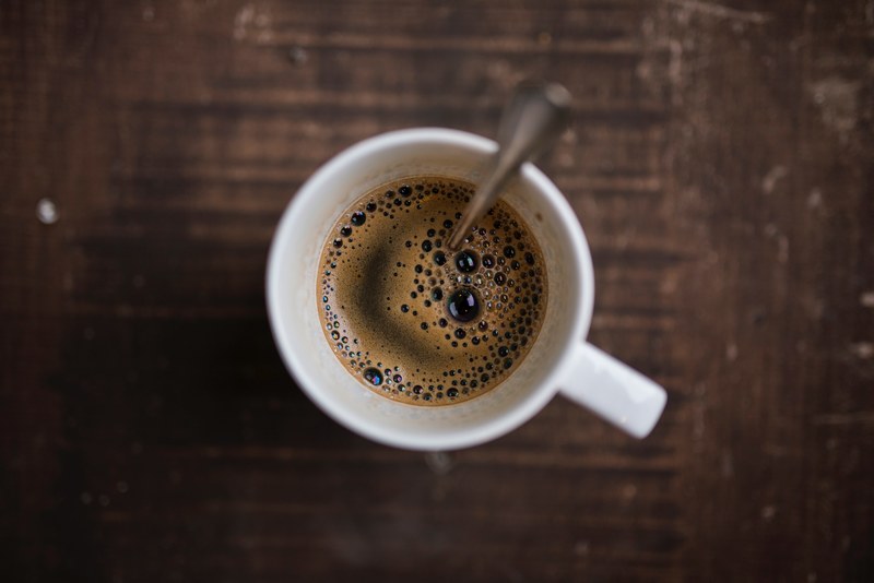 a look down into a cup of coffee in a white mug with silver spoon that sits on a dark wooden table, coffee on table with spoon 4k