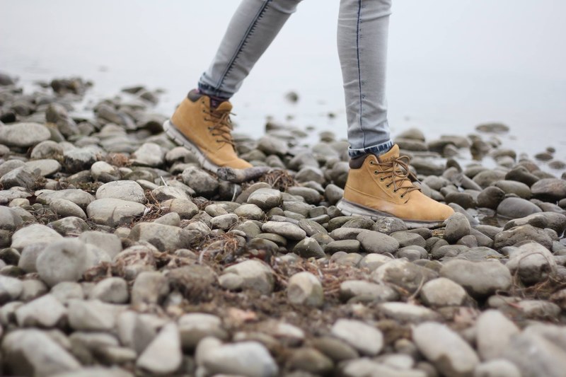 a person walking in brown boots across large pebbles by a river in rohrspitz, skipping rocks 4k