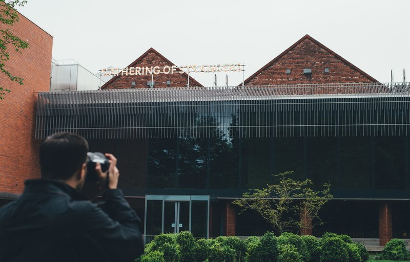 man taking picture of gathering of strangers sign on glass roof architecture at whitworth art gallery, man takes picture of brick building 4k