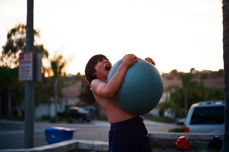 shirtless boy squeezing and playing with large inflatable ball near parked car in oceanside, shirtless boy squeezing large inflatable ball 4k