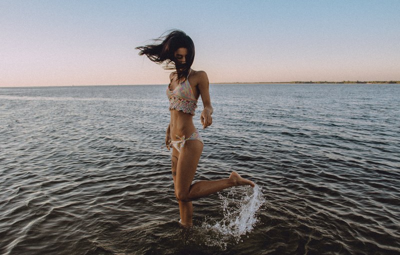 a girl in a bikini flicks her leg up behind her in the water at a beach, woman splashing water at beach 4k