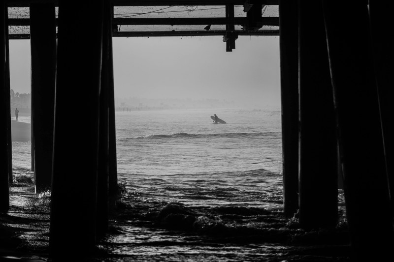 surfer with a surfboard viewed through pier pillars at santa monica state beach in black and white, surfer by the boardwalk 4k