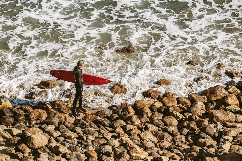 surfer with a surfboard standing on beach rocks watching the ocean at point dume state beach, surfer sizing up the waves 4k