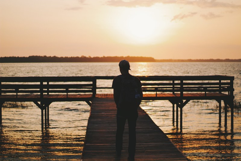 a man wearing a baseball cap on a duck at sunset in silhouette, man on dock 4k
