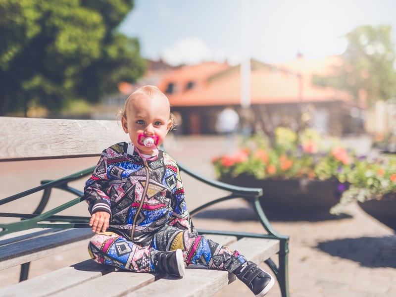 baby with a pacifer in a geometric trendy onesie on a park bench, trendy toddler clothes 4k