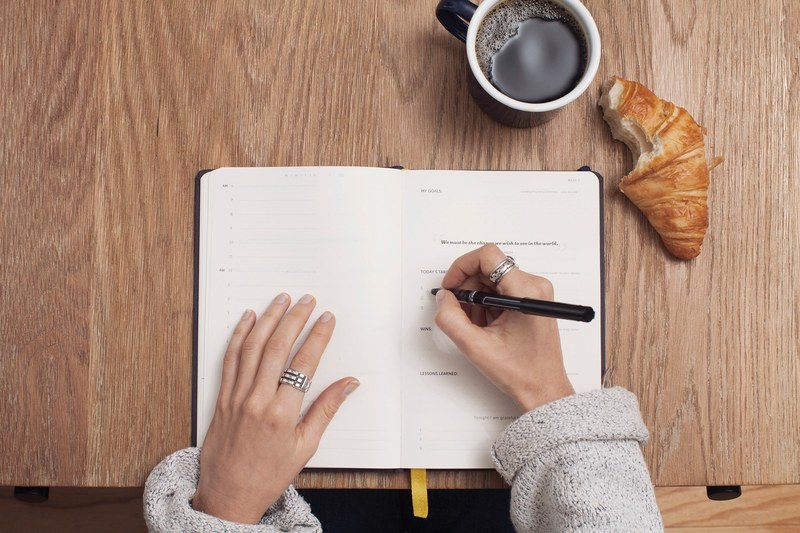 a person writing in a life planner with a coffee and a croissant on the desk, writing in a journal 4k