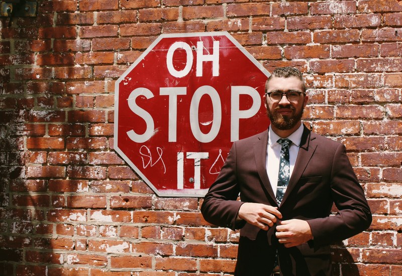 a bearded man in a brown suit in front of a brick wall with a red stop sign, oh stop it 4k