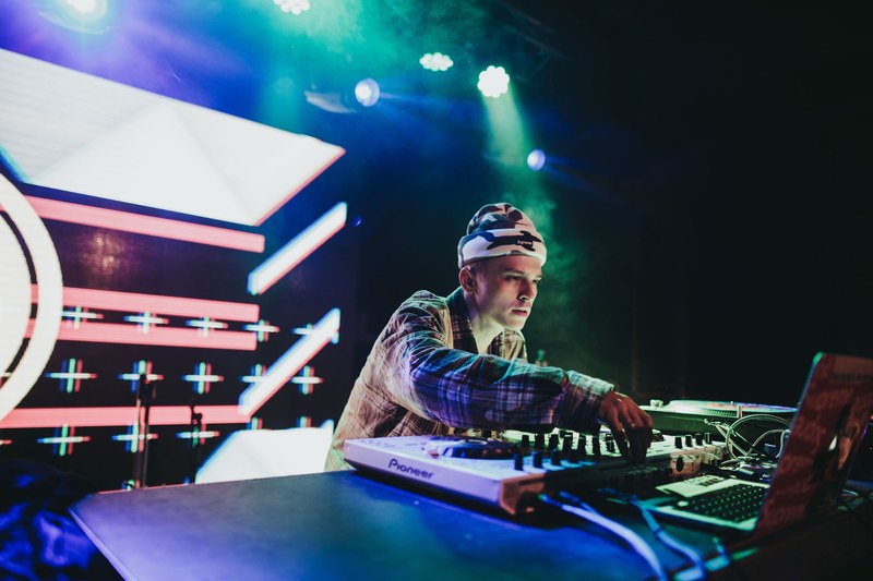 a dj in a beanie adjusting the dials on a turntable on an illuminated stage, party night 4k