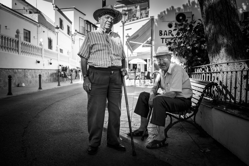 black and white photograph of two older males sitting on a bench and standing with a cane, _two old men in an ancient town in andalusia spain 4k