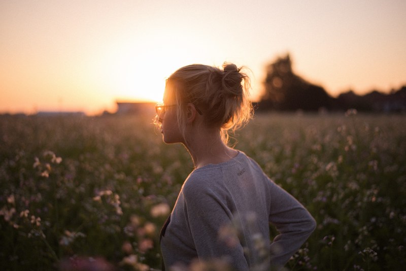 woman in a messy bun and glasses standing in a field alone at sunset, alone at golden hour 4k