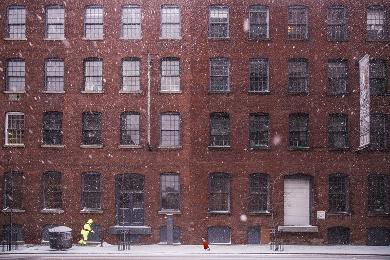 a person bundled in reflective yellow clothing shovels a sidewalk in front of a large red brick building during a snowfall, person shovels during snowfall 4k
