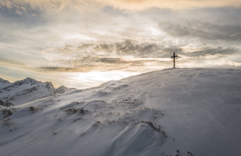a cross at the top of a snowy mountain backlit by a setting or rising sun, cross at mountain peak 4k