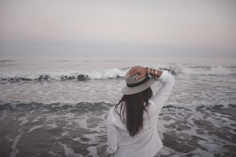 woman in white shirt holding hand on a hat watching the ocean waves on a cloudy day, woman in hat looking at ocean 4k