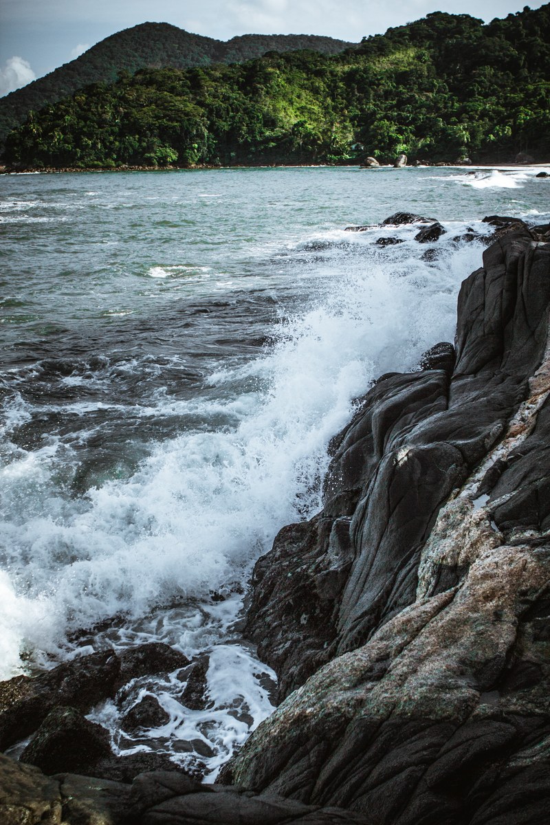 waves hitting the coastal rocks of a seashore with green mountainous land in the background, waves in front of the mountains 4k