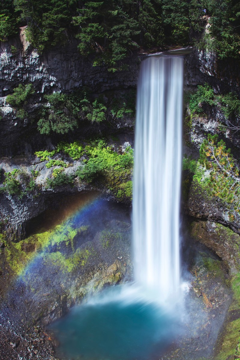 a massive waterfall flowing into a bright blue colored area with a rainbow in british columbia, smooth waterfall 4k