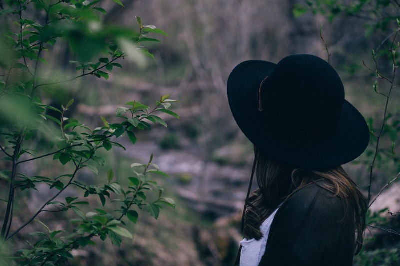 a woman in a hat standing back to camera near green branches, woman among green branches 4k