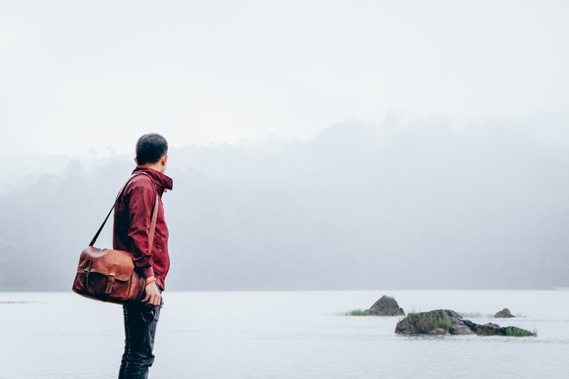 a person wearing a red jacket and brown leather bag looking out at a lake in ciwidey, foggy beach walks 4k